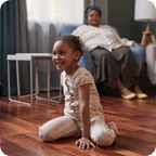 Young girl smiles and sits on the living room floor. Mother smiles at her from armchair.